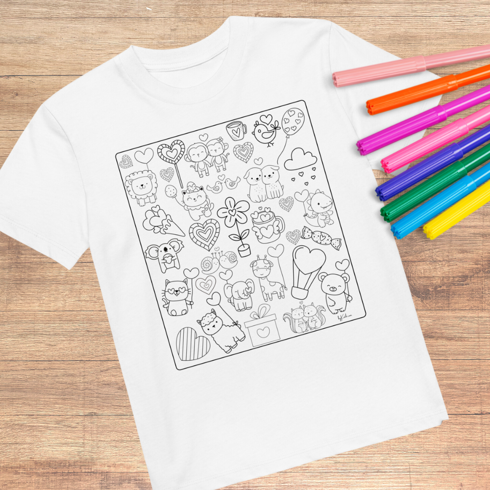 The Heart Doodles Color-in Tee, an organic cotton kids t-shirt by byKath.com, is displayed on a wooden surface featuring various cute animal and heart illustrations. Several colorful markers are placed to the right of the t-shirt, suggesting potential for coloring the drawings.