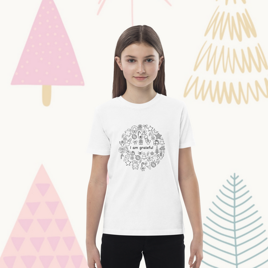 A young person with long hair stands facing forward, wearing the Gratitude Doodles Color-in Tee from byKath.com. This organic cotton kids' T-shirt features a circular pattern of playful illustrations with the phrase "I am grateful" at the center. The background showcases stylized tree designs in pastel colors.